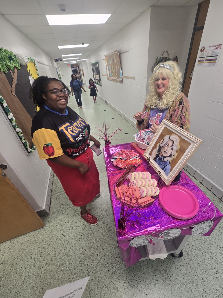 Two women in a school hallway, one in a red skirt, standing at a table with food and a picture.