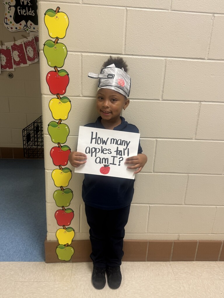 A child in a classroom holds a sign reading "How many apples tall am I?" while standing by a wall.