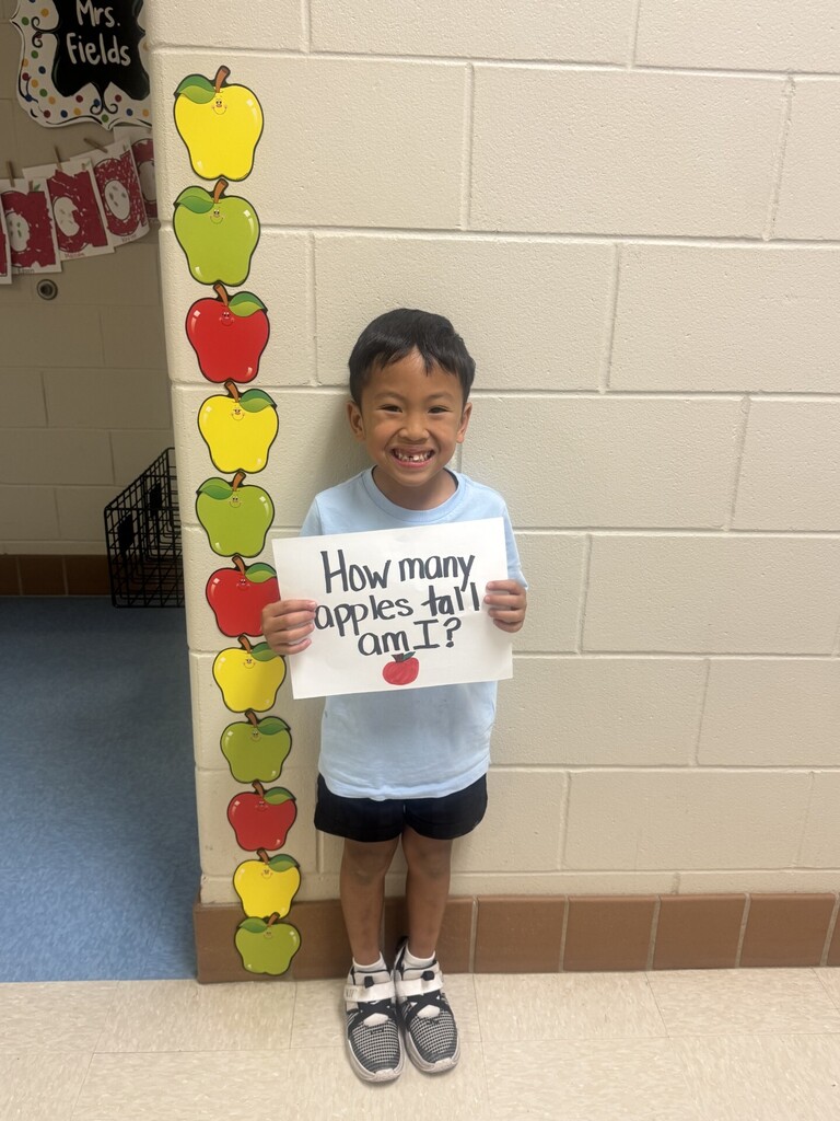Child in blue shirt and black shorts holding sign "How many apples tall am I?". Behind them, a wall with apple decorations.