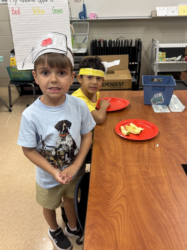 Two boys stand at a table, one in a chef's hat and another with a headband. They have plates of food in front of them.