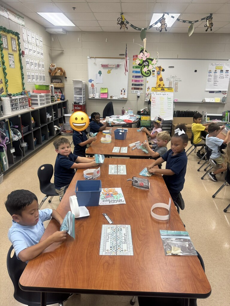 Several children seated around desks in a classroom, working on an activity. A whiteboard and various educational posters are visible.