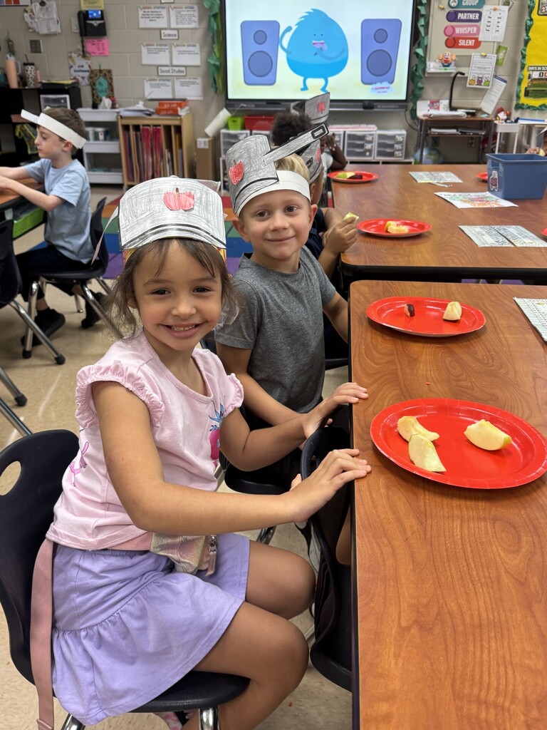 Two children in a classroom wear paper hats. One child holds a plate with slices of fruit.