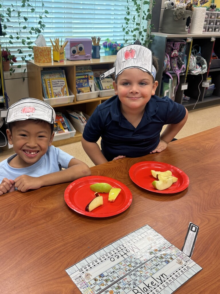 Two children in blue shirts sit at a table with red plates, one holding a paper chef's hat.