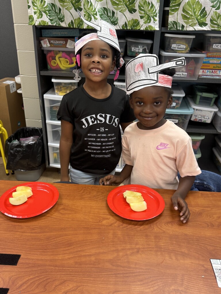 Two kids stand by a table, each wearing a paper chef hat. One holds a plate with yellow food. Behind them, shelves with storage boxes and a trash bin are visible.