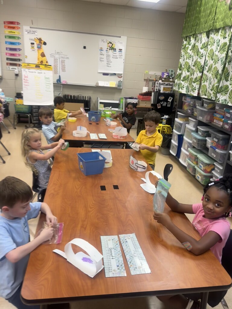 A group of children in a classroom are sitting at tables. They are doing an activity with paper and other materials.