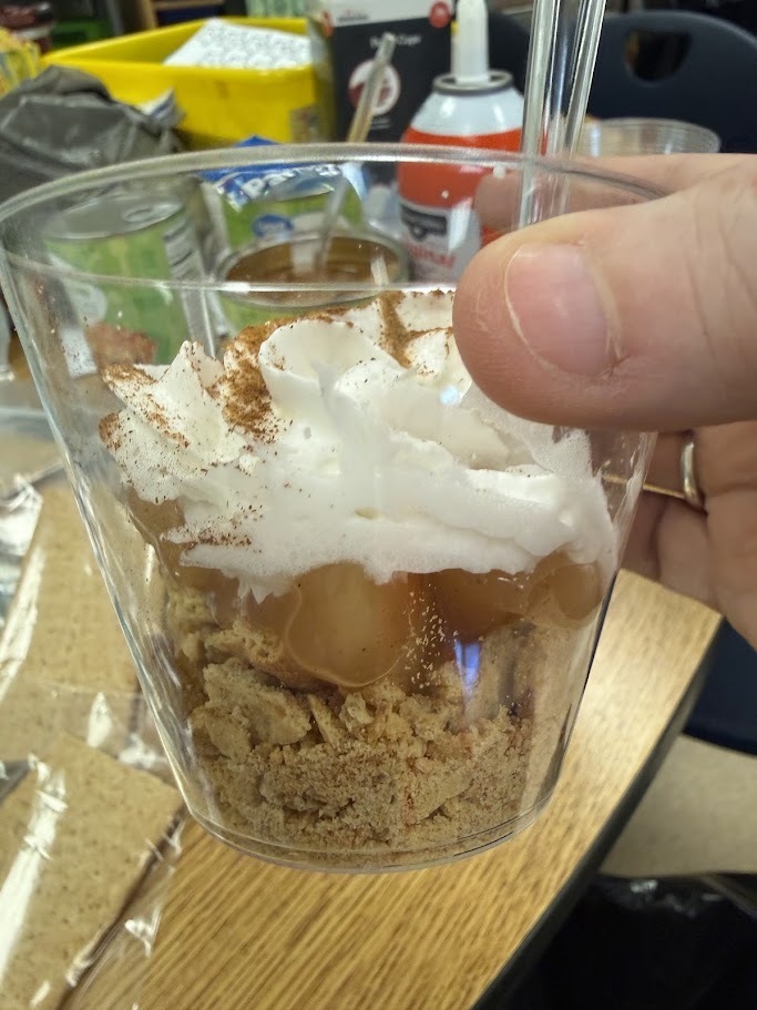 A dessert in a cup with whipped cream, caramel, and crumbly topping. A person holds the cup.