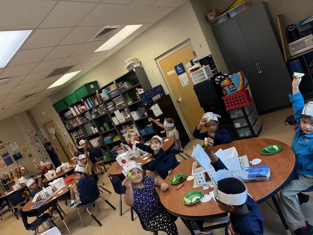 Children in a classroom wearing headbands, seated at tables, engaging in a lesson. Background includes shelves, a door, and ceiling lights.