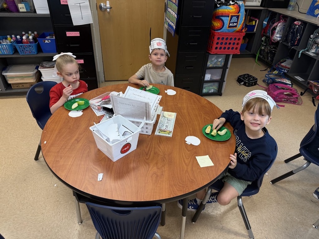 Three children sit at a round table with food and hats, smiling in a classroom.