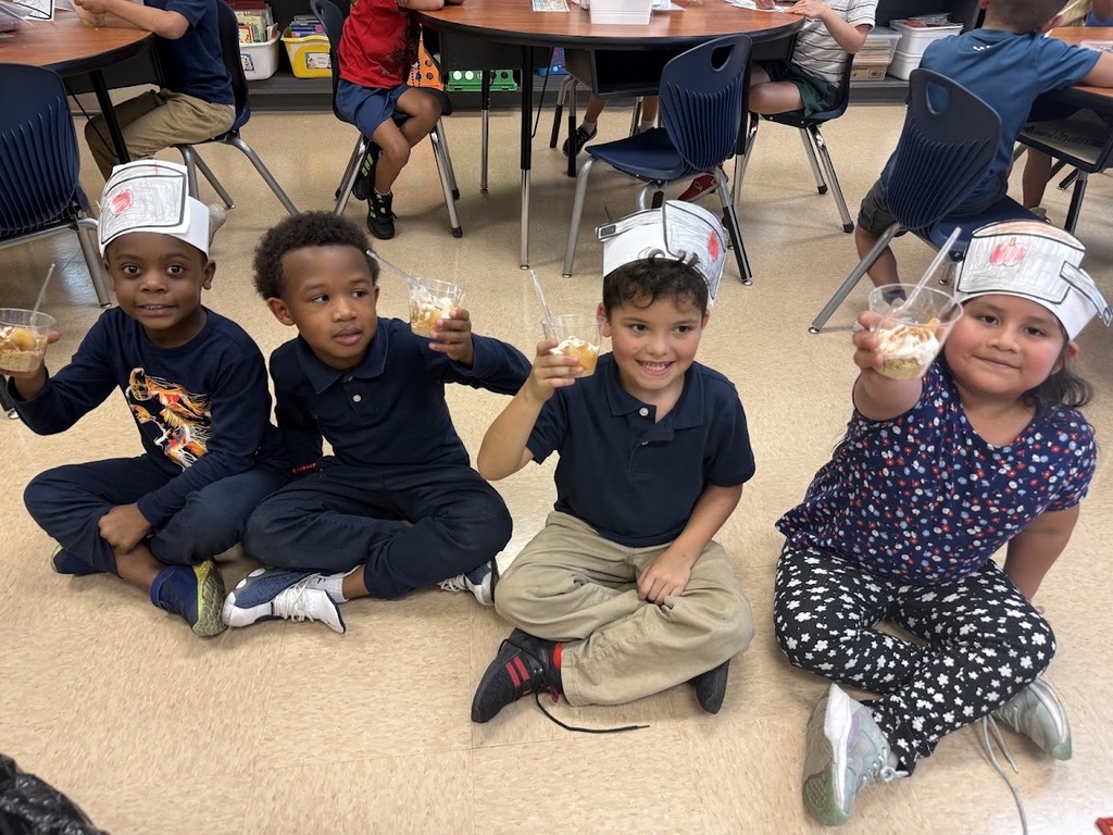 Four children sit on the floor in a classroom, wearing chef hats and holding cups. One has a dessert.