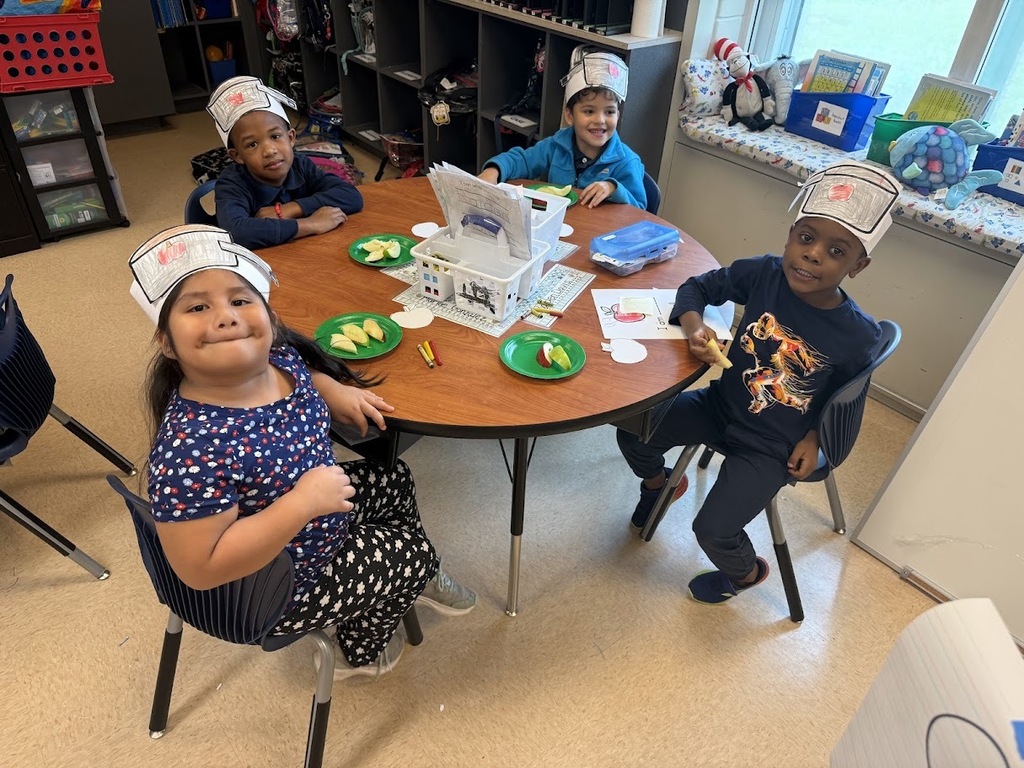 Four children wearing chef hats sit around a table with food, plates, and utensils. Background includes shelves, bins, and a window.