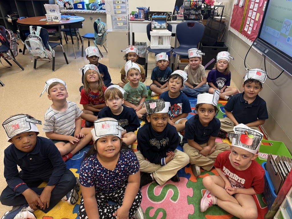 Children in a classroom wearing white hats sit on a rug, facing forward. A carpet with letters and numbers is on the floor.