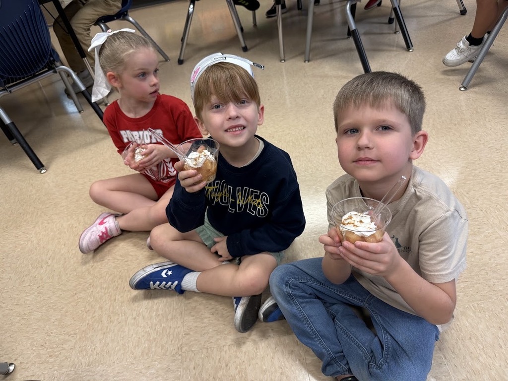 Three children sit on the floor in a classroom. One holds a dessert in a bowl, another has a dessert on a stick, and the third wears a headband.