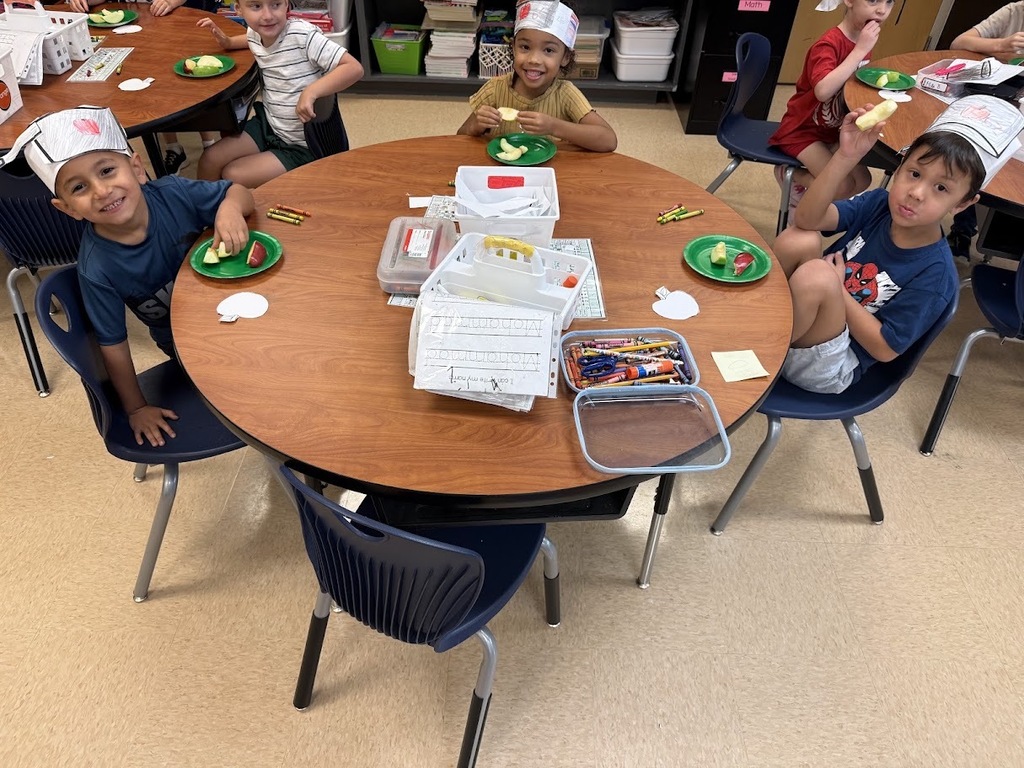 Children in chef hats sit around a table with plates of food. Some children raise their hands.
