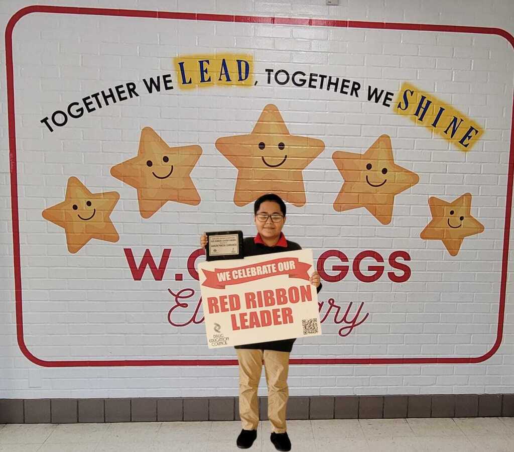 student standing in hallway holding red ribbon leader sign and plaque