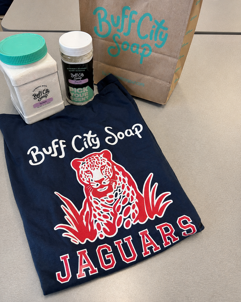 A blue shirt with "Buff City Soap" and "JAGUARS" printed, next to a brown bag, a bottle of soap, and a small bottle.