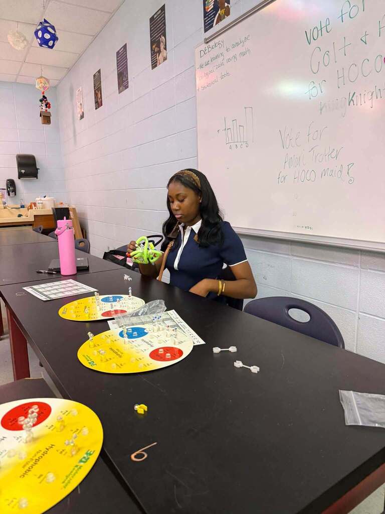 A student sits at a lab table, engaged with an experiment. Circular materials and a white board with writing are visible.