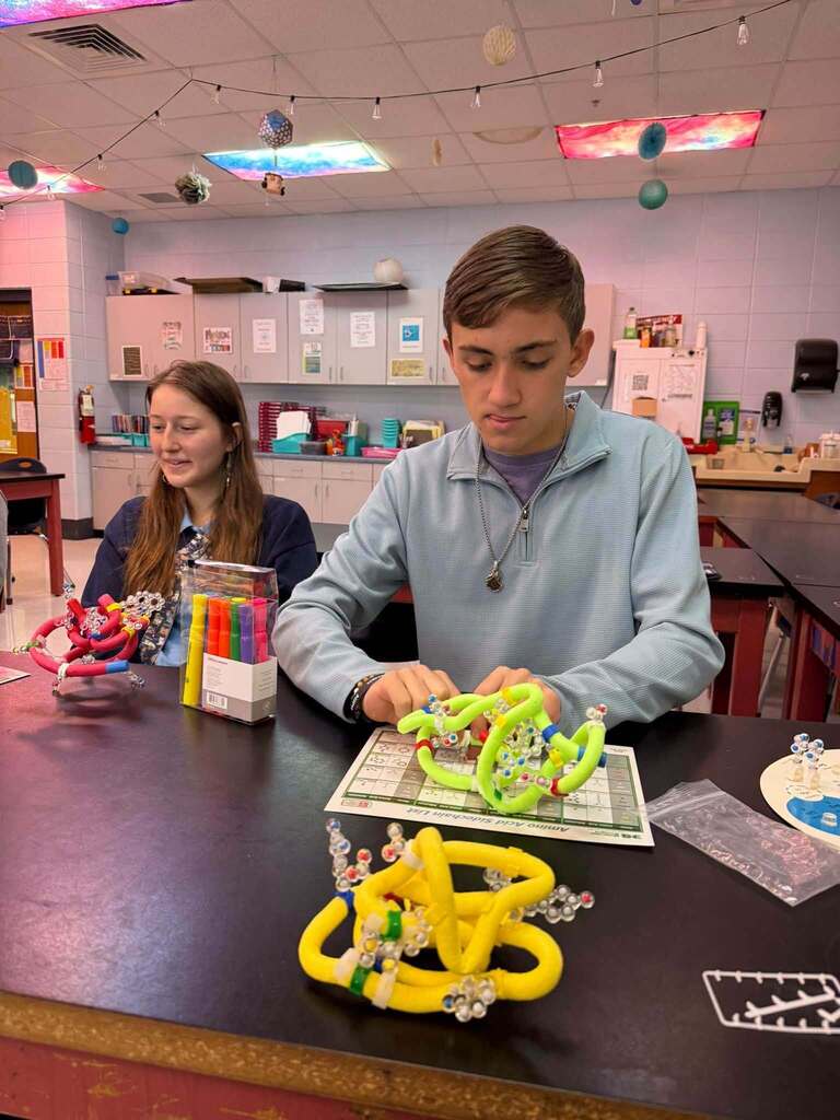 Two people sit at a table, constructing a structure from yellow and green pieces. A whiteboard, colorful objects, and a cabinet are in the background.