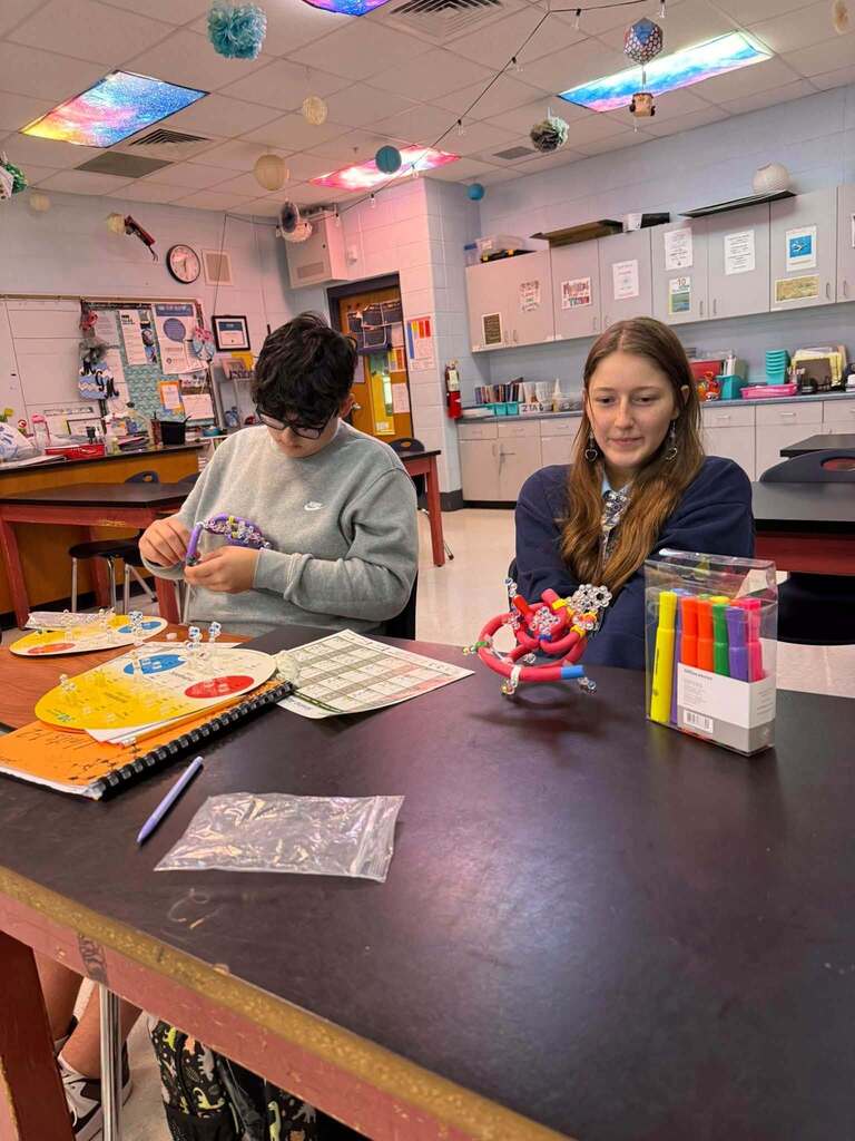 Two students seated at a table in a classroom with art supplies, a bowl, and notebooks.