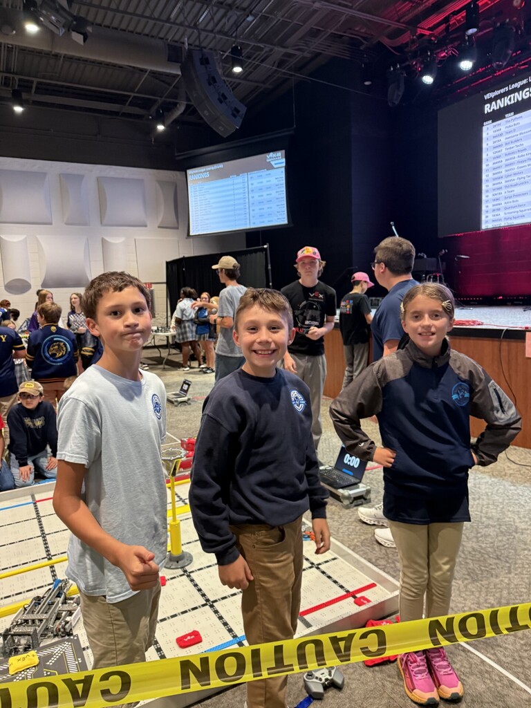 Three children stand at a table with a robot. A yellow caution tape with white letters is in front of them.