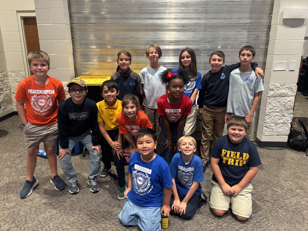 Group of kids, some wearing shirts, smiling in front of a silver wall and a yellow desk.
