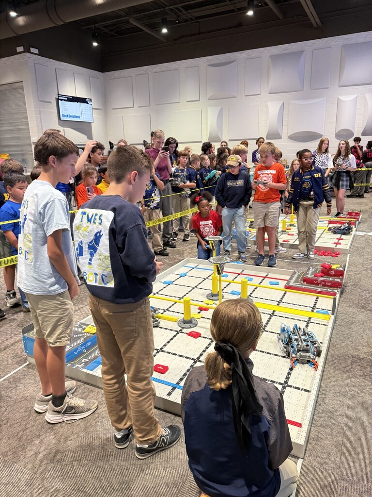 Group of children, some standing, some seated, watching a robot competition on a floor. Yellow tape marks boundaries.