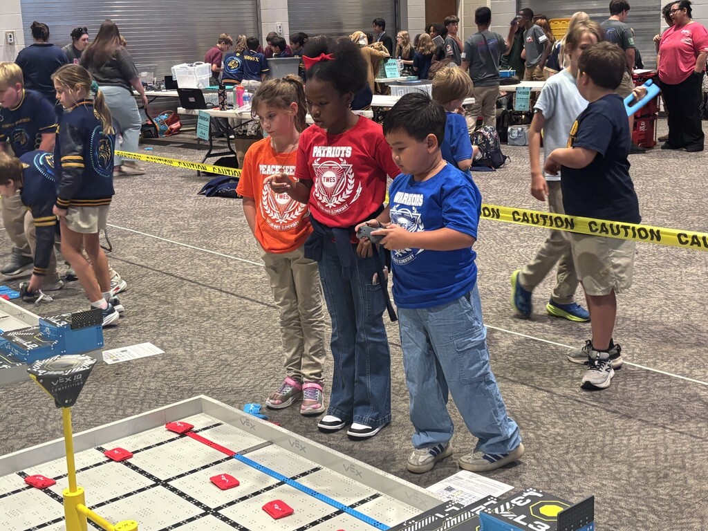 Several children observe a game on a square table with various markings and objects. They are positioned behind a yellow caution tape.