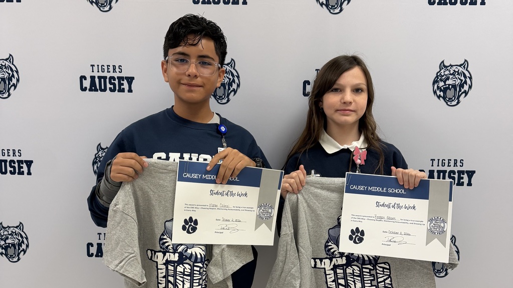 Two kids stand side by side, holding certificates, and wearing navy blue shirts in front of a wall.