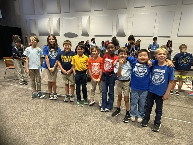 A group of children wearing various colored shirts stand close together on a carpeted floor. Some people are in the background.