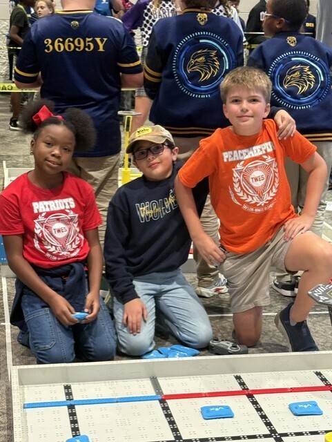 Three children wearing different jerseys, two girls and one boy, pose together in front of a game board.