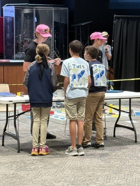 Several children wearing hats stand around a table with a white surface. A woman stands next to them, signing papers.
