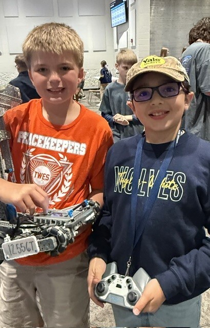 Two boys, one with an orange shirt and the other with a blue shirt, hold game controllers and smile.