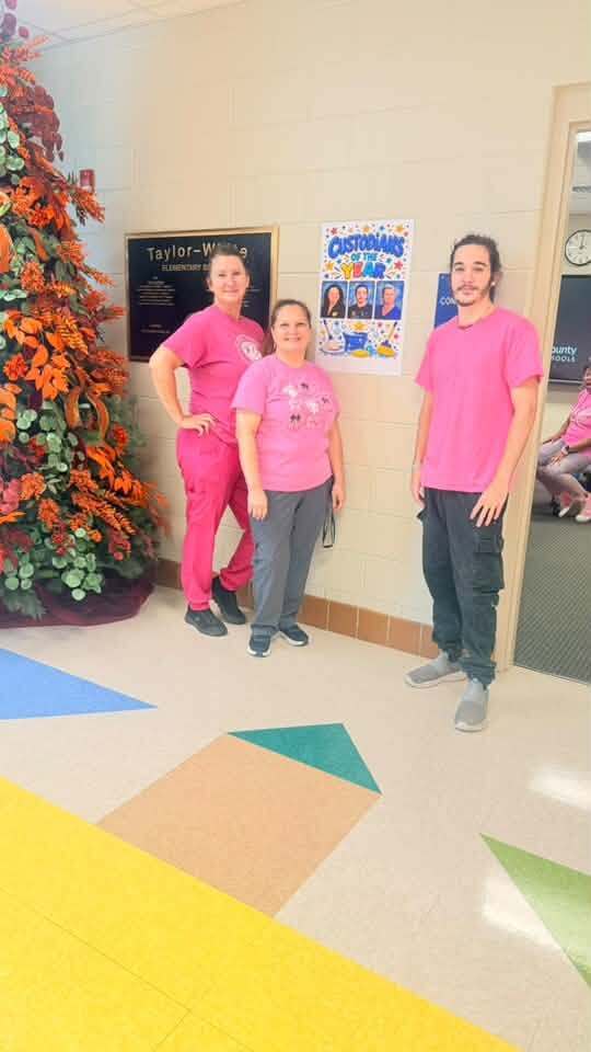 Three people stand in a corridor; two wear pink shirts. A decorative tree and bulletin board are behind them.