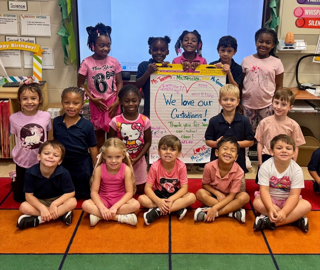 A group of young children in a classroom pose for a photo with a sign that says, "We love our Custodians."
