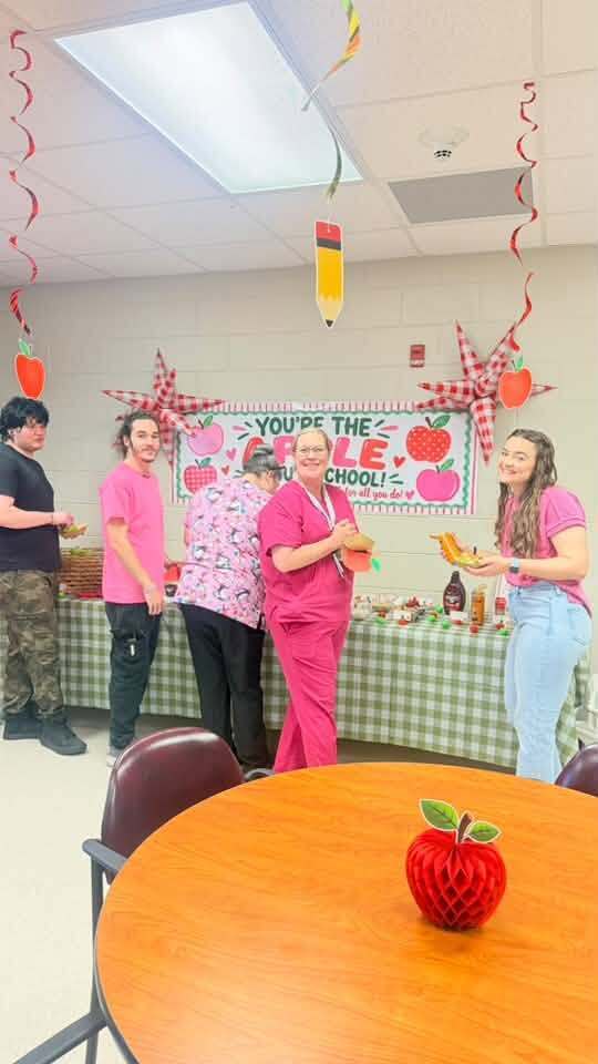 Five individuals dressed in pink uniforms gather around a table adorned with food and decorations, including apples and a banner reading "YOU'RE THE CHOOLIE."