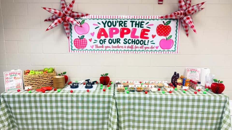 A table with a green gingham tablecloth holds various items, including apples and crafts. A decorative banner saying "You're the Apple of Our School" hangs on the wall.