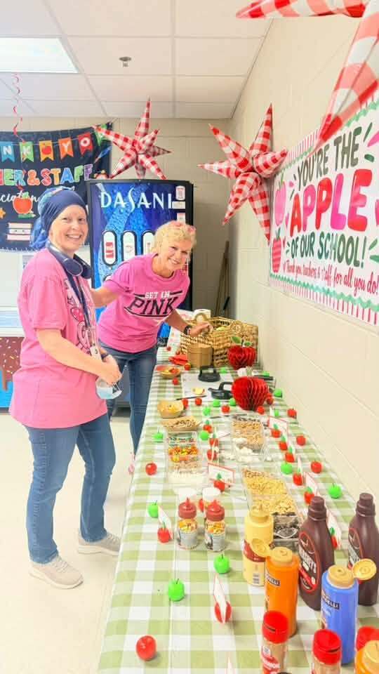 Two women in pink shirts stand by a table covered in cookies and snacks. Behind them, a banner reads "You're the apple of our school."