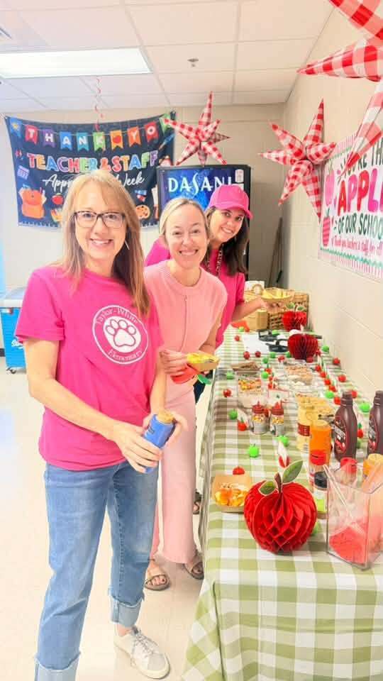 Three women in matching shirts stand by a table with food. One holds a cup, and another wears a pink hat. Behind them, a banner says "Teacher Staff".