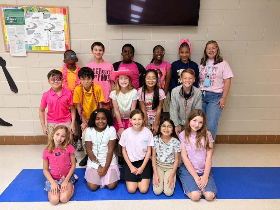 A classroom with a blue carpet displays a group of students in various poses, with some wearing pink shirts.