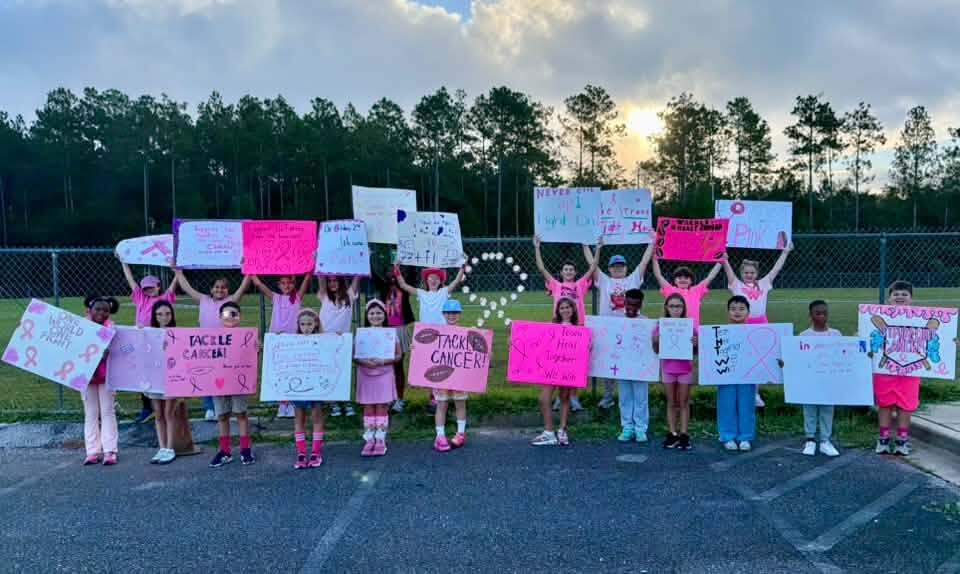A group of young people holding pink signs in a parking lot with trees in the background.