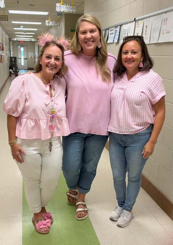 Three women wearing pink tops and bottoms stand together in a hallway. The middle woman has long blonde hair. Behind them are bulletin boards and ceiling lights.