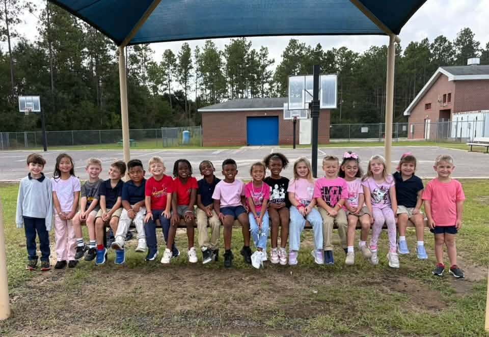 Children in various colors of shirts and pants stand and sit together under a blue awning.