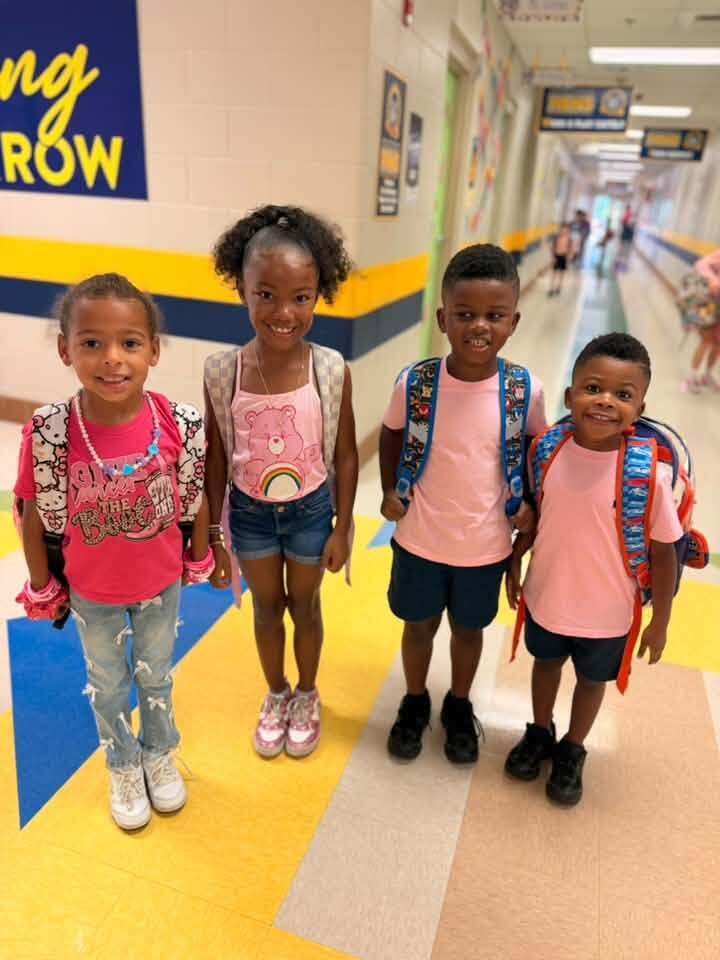Four kids stand together in a hallway. Each wears backpacks and smiles at the camera. One girl wears a pink shirt with a heart design.