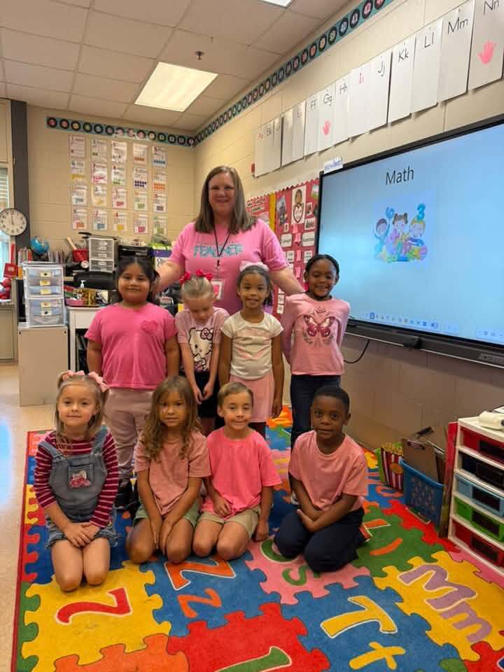 A teacher and seven children in pink shirts pose in front of a classroom screen with a math lesson.