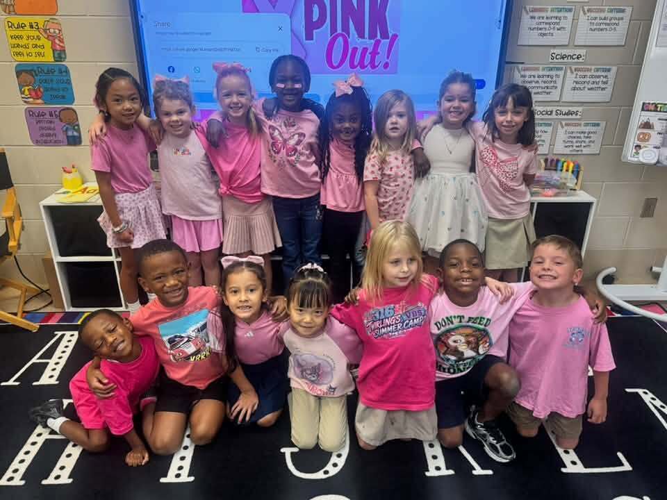 Group of young children in pink shirts pose for a photo in a classroom. They are standing on a floor with letters.