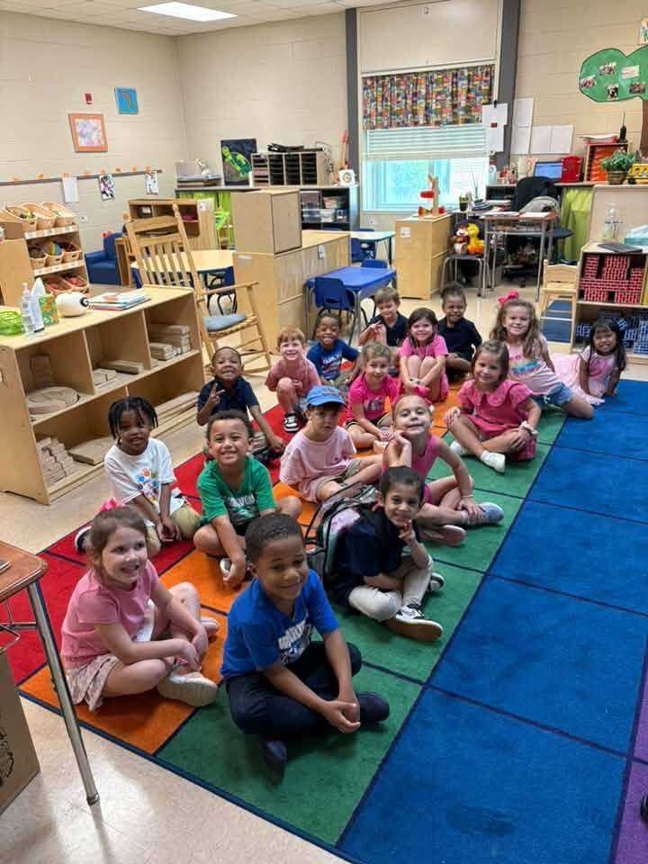 Classroom with children sitting on the floor. Some are wearing hats, sneakers, and shoes.