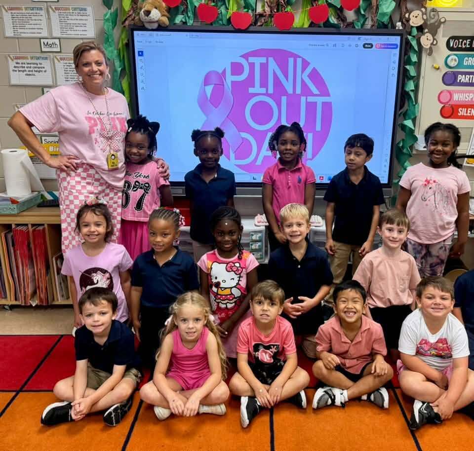Group of children, including girls in pink and boys in blue and black, pose for a photo in a classroom.