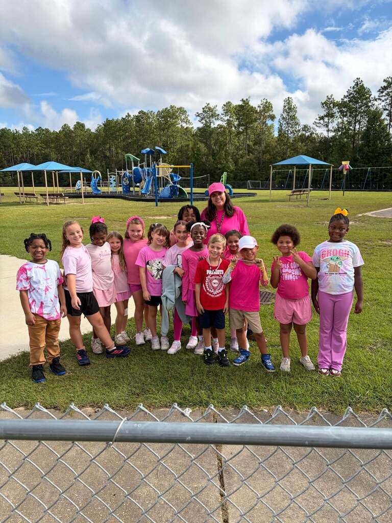 Group of young children wearing pink shirts pose for a photo in a park with playground equipment.