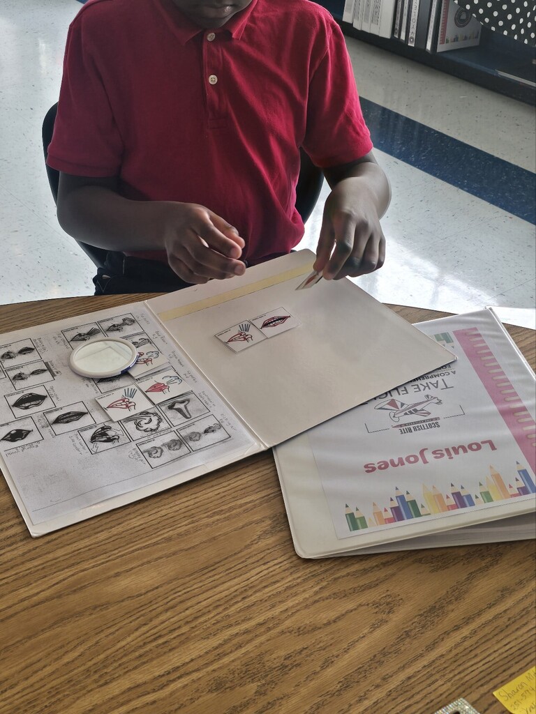 A child in a red shirt works on a worksheet with images and words on a table.