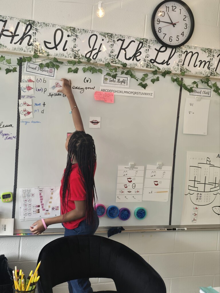 A girl in a red shirt touches a whiteboard with alphabet flashcards. A clock and vines are above the board.