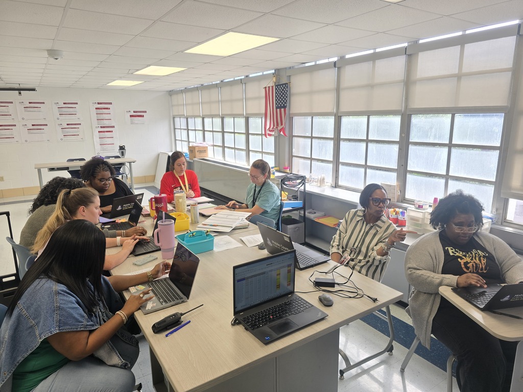 Several people in a classroom setting, seated at desks, using laptops. A large window with blinds is in the background.